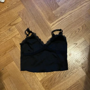 Svart ribbad linnetopp med spets - Säljer en svart croppad linnetopp med tunna justerbara axelband och snygga spetsdetaljer längs urringningen. Toppen har ribbad struktur och omlott-design framtill, vilket ger en trendig och feminin look. Perfekt att styla med jeans eller kjol.