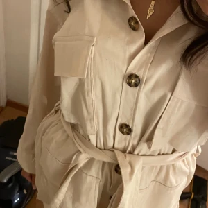 Beige Trenchcoat med bälte -  beige trenchcoat med stora bruna knappar och praktiska fickor framtill. jackan har ett matchande bälte i midjan som går att ta bort, ger en avslappnad siluett. långärmad och med klassisk krage, perfekt för dig som gillar minimalistisk stil. har slit på sidan. använd 2 ggr, skriv för fler bilder🥰