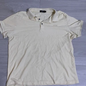 Beige pikétröja från BOSS XL - Klassisk beige pikétröja från BOSS i storlek XL. Tröjan har korta ärmar, krage med två knappar och diskret BOSS-brodyr på bröstet. Tillverkad i bomull för en skön och luftig känsla. Perfekt till jeans eller shorts för en clean look.