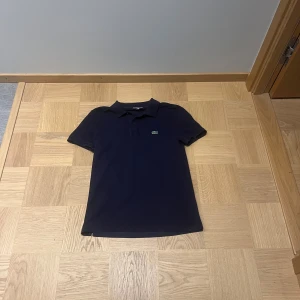 Mörkblå pikétröja från Lacoste - Snygg mörkblå pikétröja från Lacoste. Riktigt fin skick. Inga defekter. Kan posta samma dag som köpet.
