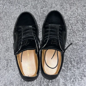 Svarta sneakers från Giuseppe Zanotti - Säljer ett par svarta sneakers från Giuseppe Zanotti med snygg platt sula och dragkedjedetaljer på sidorna. Skorna är i skinn och har rund tå samt snörning framtill. Perfekta för dig som gillar stilrena och exklusiva sneakers.