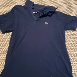 Marinblå pikétröja från Lacoste - Säljer en klassisk marinblå pikétröja från Lacoste med den ikoniska gröna krokodilloggan på bröstet. Tröjan har korta ärmar, krage och knappar vid halsen. Perfekt för en clean och sportig look.