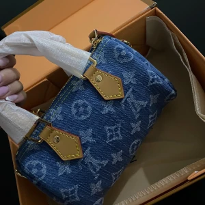 Louis Vuitton denim handväska - Superfin handväska från Louis Vuitton i blått denimtyg med ikoniskt monogrammönster. Väskan har detaljer i ljust skinn, guldfärgade metalldelar och dragkedja upptill. Kommer med både handtag och axelrem för olika stilar. Perfekt för dig som vill sticka ut med en lyxig touch.