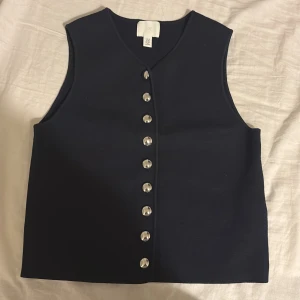 Mörkblå väst med silvriga knappar H&M - Elegant mörkblå väst från H&M med raka linjer och v-ringad hals. Västen har en rad silvriga knappar framtill som ger en cool touch. Materialet är mjukt och känns som polyester. Perfekt att styla över en t-shirt eller skjorta för en trendig look. Helt oanvänd.