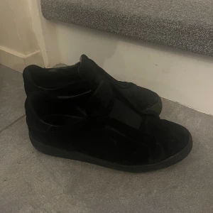 Svarta sneakers arigato - Svarta sneakers från Axel Arigato i mocka med kardborreband istället för klassisk snörning. Snygg och stilren design med platt sula och rund tå. Perfekta för dig som gillar minimalistiska och bekväma skor.