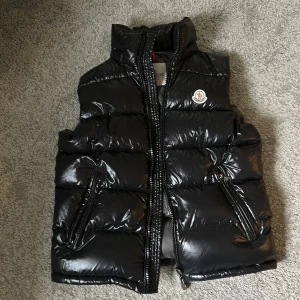 Svart dunväst från Moncler - Snygg svart dunväst från Moncler med glansig finish och hög krage. Västen har två praktiska fickor med dragkedja och ikonisk Moncler-logga på bröstet. Perfekt att slänga över en hoodie för extra värme under kyliga dagar. Aldrig använd.  OBS Storlek är 1 och inte L