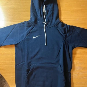 Mörkblå Nike hoodie med huva - Snygg mörkblå hoodie från Nike med vit logga på bröstet och klassisk känguruficka. Tröjan har huva med vit dragsko och långa ärmar. Perfekt för chill eller träning.