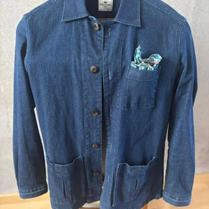 Mörkblå jeansjacka från John Henric - Snygg mörkblå jeansjacka från John Henric med klassisk krage, knäppning framtill och tre praktiska fickor. Jackan har en rak passform och är tillverkad i slitstarkt jeanstyg. Perfekt för dig som gillar en stilren och avslappnad look.