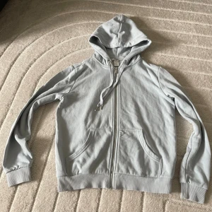 Ljusgrå hoodie från H&M med dragkedja - Säljer en ljusgrå hoodie från H&M i storlek XS. Tröjan har huva med snörning, dragkedja framtill och två stora fickor. Tillverkad i mjuk bomullsmix och har ribbade muddar vid ärmslut och nederkant. Perfekt för chill dagar eller när du vill vara extra bekväm.