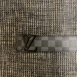 Louis Vuitton Damier Graphite bälte - Snyggt Louis Vuitton bälte i svart skinn med klassiskt Damier Graphite-rutmönster och en stilren LV-logga i metall som spänne. Bältet är tillverkat i Spanien och har en exklusiv känsla som lyfter vilken outfit som helst.