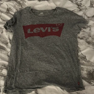 Grå Levi's t-shirt med röd logga - Snygg grå t-shirt från Levi's med klassisk röd logga framtill. T-shirten är i mjuk bomull och har en normal passform med korta ärmar. Perfekt för en avslappnad och cool stil.