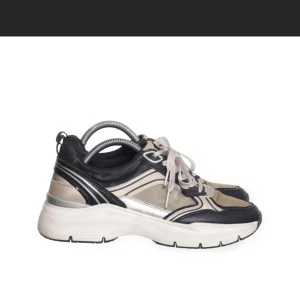 Tamaris  sneakers i svart och beige - Säljer ett par Tamarissneakers med cool retrovibe i svart, beige och silver. Skorna har platt sula, rund tå och snörning framtill. Materialmix med syntet och skinnliknande detaljer ger en schysst kontrast. Perfekt för dig som gillar streetstyle och vill sticka ut.