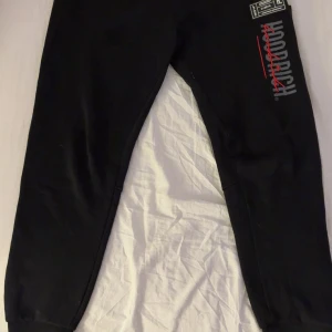 Svarta mjukisbyxor från Hoodrich  - Svarta mjukisbyxor från Hoodrich i storlek medium. Byxorna har tryck i vitt och rött på benet och detaljerade loggor. De är tillverkade i mjukt material med resår i midjan och muddar vid bensluten för en skön passform.