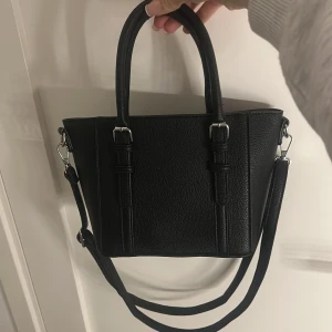 Svart handväska med axelrem - Stilren svart handväska i skinnimitation med både handtag och avtagbar axelrem. Väskan har en klassisk, fyrkantig form och silverfärgade metalldetaljer. Insidan har två fack med dragkedja och extra fickor för småsaker. Pris kan diskuteras.