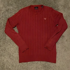 Röd kabelstickad tröja från GANT - Säljer en röd kabelstickad tröja från GANT med broderad logga på bröstet. Tröjan har rund halsringning, ribbade muddar och är tillverkad i mjuk bomull. Perfekt för dig som gillar klassisk och stilren look. Den är storlek M men den är stretch och den sitter perfekt på en som är 170! Pris kan diskuteras vid snabb affär!