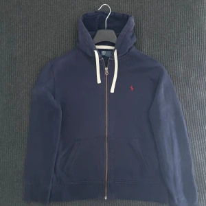 Marinblå hoodie från Polo Ralph Lauren - Snygg marinblå hoodie från Polo Ralph Lauren med dragkedja och vit snörning i huvan. Klassisk röd logga broderad på bröstet och två fickor framtill. Perfekt för dig som gillar stilren och sportig look. L/M i storlek