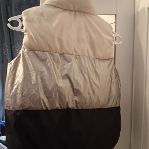Färgglad dunväst från Sinsay 140 cm - Säljer en dunväst från Sinsay i storlek 140 cm. Västen har coola blockfärger i beige, silver och svart, med hög krage och dragkedja framtill. Två fickor på sidorna gör den både snygg och praktisk. Perfekt för att ge din outfit lite edge.