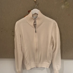 Ljus Beige kashmirtröja från Gran Sasso - Exklusiv beige tröja från Gran Sasso i 100% kashmir. Tröjan har ribbad hög krage, hel dragkedja framtill och ribbade muddar vid ärmslut och midja. Perfekt för dig som gillar stilrena och lyxiga plagg med en clean look. Knappt använd. Tyvärr har den två små hål på vänstra ärmen men de är väldigt svåra att märka. Den har även två små fläckar längst ned som är svåra att se men kan skicka bild om man vill se tydligare.