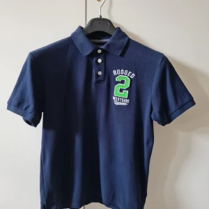 Pikétröja med tryck från Cubus strl 164 (barn) - Säljer en mörkblå pikétröja med krage och tre knappar framtill från Cubus. På bröstet finns ett broderat tryck med texten 'Rugged 2 Westside'. Material 100% bomull. Storlek 164. Kan hämtas i Vallentuna.