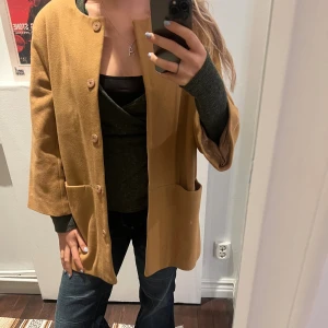 Beige kappa från Stylein i ull - Säljer en beige kappa från Stylein i storlek S. Jackan har rund hals, stora fickor framtill och stängs med knappar. Den är tillverkad i mjuk ull och har en clean, rak passform med långa ärmar. Perfekt för dig som gillar minimalistisk och stilren vibe.