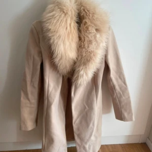 Beige kappa med fluffig päls - Snygg beige kappa med stor fluffig krage i ljus päls. Jackan har en rak och stilren form med långa ärmar och är tillverkad i mjukt material. Perfekt för dig som vill ha en classy och trendig look under höst och vinter. Storlek M 