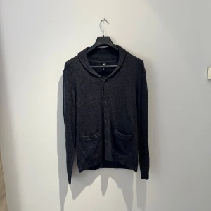 Mörkgrå kofta från H&M med knappar - Mysig mörkgrå kofta från H&M i storlek M. Koftan har långa ärmar, två framfickor och stängs med knappar framtill. Den har en mjuk krage och ribbade muddar vid ärmslut och nederkant. Perfekt för lager-på-lager under kyliga dagar. Hör av er vid frågor eller fler bilder