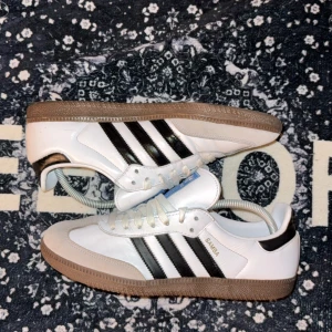 Adidas Samba sneakers vit/svart - Klassiska Adidas Samba sneakers i vitt skinn med svarta ränder och detaljer. Skorna har brun gummisula, snörning och rund tå. Märket syns tydligt på tungan och sidan. Perfekta för dig som gillar retrostil och streetwear.väldigt fina knappt använda 