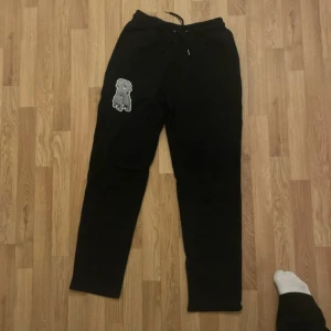 Svarta mjukisbyxor från risky med patch - Svarta mjukisbyxor från SRY i storlek M. Byxorna har en elastisk midja med snörning och en grå patch med motiv på vänster lår. Perfekta för chill dagar eller när du vill ha det bekvämt. Mjukt material och avslappnad passform.