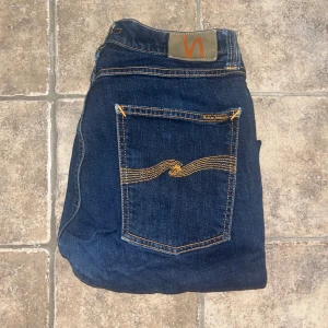 Nudie jeans  - Snygga mörkblå jeans från Nudie med klassisk orange söm på bakfickorna och logga på linningen. Midja 36cm, längd 91cm (möjligen uppsydda då jag tycker de är mer som L30) lagad i grenen (se bild ) och gylfen fungerar ej! JAG SKICKAR EJ FLER BILDER NÄR JAG BÄR JEANSEN! för storleksguide hänvisar jag till måtten och för bättre inblick i passformen rekommenderar jag att googla på modellnamnet. :) S4
