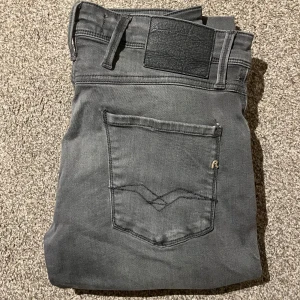 Replay Anbass grå jeans, storlek 29 - Snygga Replay Anbass jeans i grått med klassisk femficksdesign och svarta detaljer på bakfickan. Jeansen har en slim passform och är tillverkade i mjukt bomullsmaterial med stretch för extra komfort. Perfekt för dig som gillar stilrena och moderna jeans.