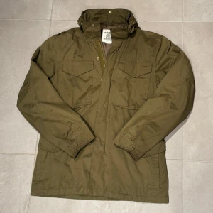 Olivgrön feild jacka från Dobber - Säljer en olivgrön field jacka från Dobber med hög krage och dragkedja framtill. Jackan har flera praktiska fickor med lock och en klassisk, rak passform. Perfekt för dig som gillar stilren och funktionell design.