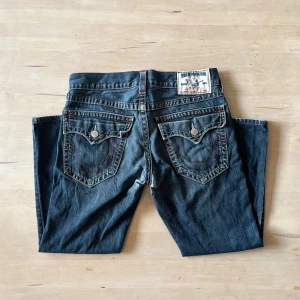 True Religion jeans med röd söm, stl 32 - Snygga blå jeans från True Religion med ikoniska röda sömmar och detaljerade bakfickor med lock och knapp. Klassisk femficksmodell, tillverkade i USA. Jeansen har en rak passform och snygga slitningar. Lite slitna i grenen och har lite heel bite, syns på bilderna.