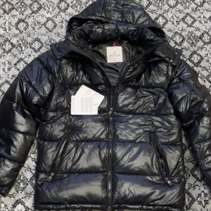 Svart Moncler pufferjacka med huva - Svart pufferjacka från Moncler med glansig finish, huva och dragkedja framtill. Jackan har två snedställda fickor med dragkedja, quiltad design och Moncler-logga på ärmen. Insidan har en cool serietidningspatch. Perfekt för kalla dagar.