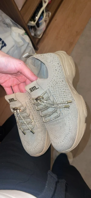 Beige glittriga sneakers från STL - Snygga beige sneakers från STL med stickad ovandel och glittriga snören. Skorna har en chunky platt sula och detaljer av små glittrande stenar över hela ovandelen. Perfekt för dig som gillar att sticka ut med stilrena och trendiga skor.