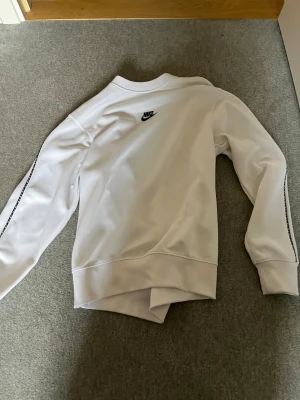 Vit Nike sweatshirt - Säljer en fräsch vit sweatshirt från Nike i storlek XS. Tröjan har svart Nike-logga på bröstet och svarta detaljer längs ärmarna. Materialet är mjukt och känns sportigt, perfekt för chill eller träning. Rund hals och långa ärmar ger en clean look.