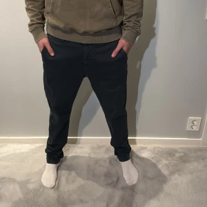 Mörkblå Replay chinos med tapered fit - Snygga mörkblå chinos från Replay med tapered passform och klassiska fickor både fram och bak. Byxorna har bälteshällor och subtil Replay-logga ovanför bakfickan. Materialet är mjukt och har en lätt glansig finish som ger en modern vibe. Modelllen är 186cm och väger 70kg 