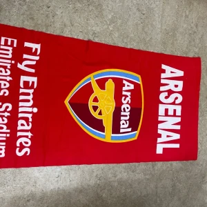 Röd Arsenal halsduk Emirates Stadium - Stor röd halsduk med Arsenal-logga och texten 'Fly Emirates Emirates Stadium'. Tydliga vita bokstäver och klubbens klassiska kanon i gult och blått. Perfekt accessoar för alla Arsenal-fans som vill visa sitt stöd på matchdag.