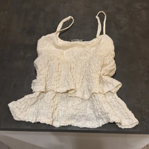 Krämvit volangtop från Gina Tricot - Säljer en krämvit linnetopp från Gina Tricot med smala axelband och snygga volangdetaljer. Toppen har en skrynklig textur som ger en cool och trendig look. Perfekt för dig som gillar romantiska vibes och vill sticka ut lite extra.💞💞
