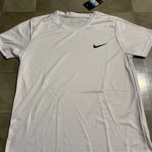 Vit Nike Dri-FIT t-shirt herr M - Fräsch vit t-shirt från Nike med svart swoosh-logga på bröstet. Tillverkad i lätt och ventilerande Dri-FIT polyester, perfekt för träning. Klassisk rund hals och korta ärmar. Snygg och sportig stil som passar dig som gillar att hålla dig aktiv.