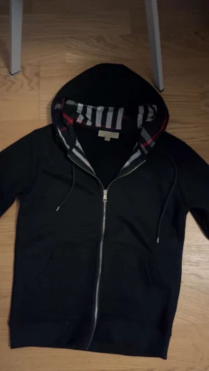  Burberry hoodie - Svart hoodie från Burberry med klassiskt rutigt foder i huvan och snygga silverdetaljer på dragkedja och snörning. Tröjan har hel dragkedja framtill, fickor och långa ärmar. Perfekt för dig som vill ha en stilren och lyxig look.