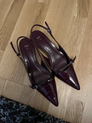 Vinröda pumps med spetsig tå från Zara - Snygga vinröda pumps från Zara i storlek 39. Skorna har en glansig finish, spetsig tå och smal rem med spänne över foten. Liten klack och öppen häl ger en elegant vibe. Perfekta för dig som gillar stilrena och trendiga skor.