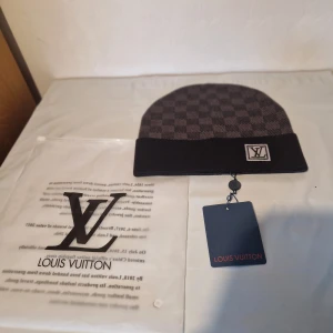 Louis Vuitton rutig mössa i ull - Snygg mörkgrå och svart mössa från Louis Vuitton med klassiskt rutigt mönster och LV-logga framtill. Tillverkad i mjuk ull som håller dig varm och stilren under kyliga dagar. Perfekt accessoar för dig som gillar exklusiva märken.