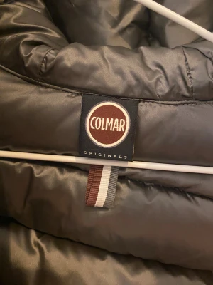 Svart Colmar pufferjacka med huva - Säljer en svart pufferjacka från Colmar med stor, mysig huva och markerad logga på ärmen. Jackan har dragkedja framtill, två fickor med dragkedja. Den är väldigt varm och fin. Har dock inte kommit till användning så den är nästan helt ny 