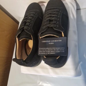 Svarta sneakers från Christian Louboutin 44 - Snygga svarta sneakers från Christian Louboutin med signaturbroderi på hälen och klassisk röd sula. Skorna har snörning och är tillverkade i mjuk mocka med detaljer i skinn på insidan. Perfekt för dig som gillar exklusiv och stilren design.