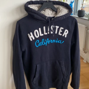 Mörkblå Hollister hoodie med foder - Mysig mörkblå hoodie från Hollister med stor vit och blå broderad text på bröstet. Insidan av huvan är fodrad med fluffigt foder, och tröjan har känguruficka samt snörning vid huvan. Perfekt för kalla dagar och chill stunder.