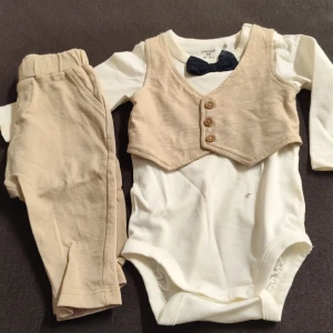 Söt babykostym med väst och fluga Lindex - Supersöt babykostym från Lindex i storlek 62. Setet består av en cremevit långärmad body med fastsydd beige väst med knappar och en mörkblå fluga. Tillhörande byxor i matchande beige färg. Perfekt för speciella tillfällen!