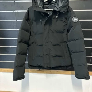 Svart pufferjacka från Canada Goose - Svart pufferjacka från Canada Goose McMillan modellen med huva och snygg svart patch på ärmen. Jackan har dragkedja och knappar framtill, samt två praktiska fickor. Perfekt för kalla dagar med sin tjocka, quiltade design och stilrena look.