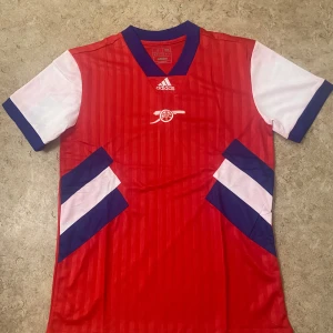 Arsenal röd fotbollströja Adidas - Säljer en snygg Arsenal fotbollströja från Adidas i rött med vita korta ärmar och blå detaljer. Tröjan har ett subtilt randigt mönster på framsidan, blå V-ringad krage och vita/blå grafiska inslag på sidorna. Tillverkad i lätt och ventilerande polyester.