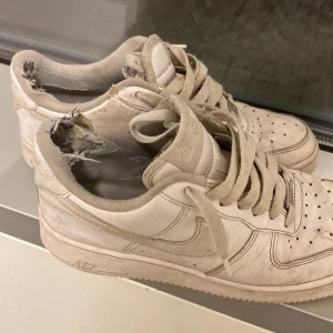 Nike Air Force 1 vita sneakers - Nike Air Force 1 sneakers i vitt skinn med klassisk låg siluett och rund tå. Skorna har perforerad tåbox, snörning och platt sula. Swoosh-logga på sidan och AIR-detalj på hälen. Perfekt för dig som gillar retro streetstyle.