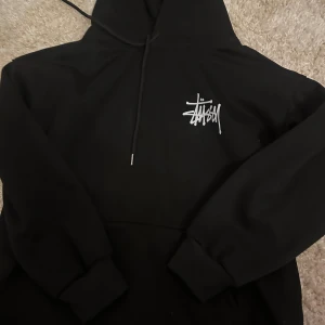 Svart Stüssy hoodie - Svart hoodie från Stüssy i storlek M med vit logga på bröstet och stort grafiskt tryck på ryggen. Tröjan har huva med snörning och ribbade muddar. Bra skick har använts få gånger 